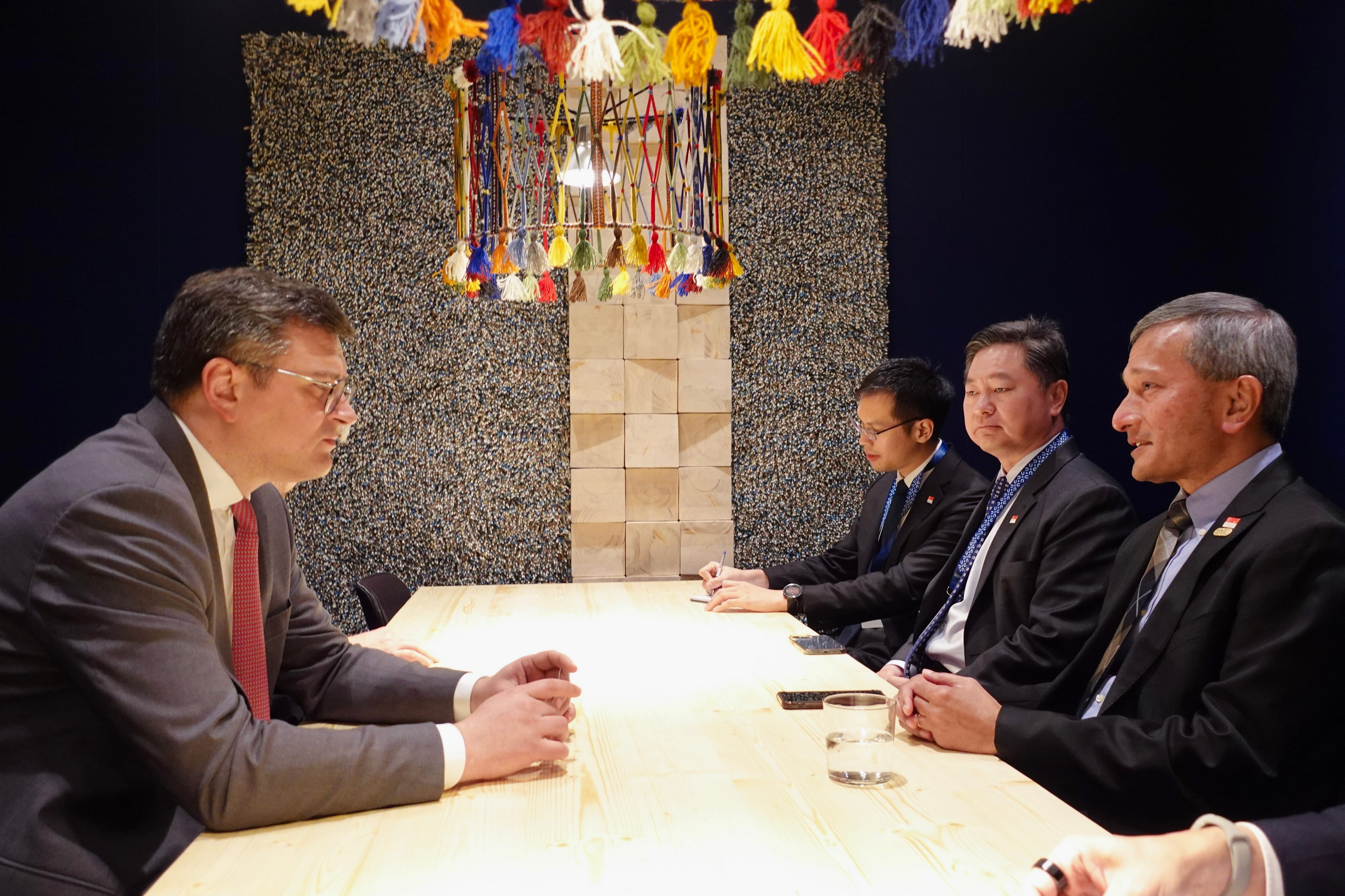 Four men in suits sit at a light wood table under a colorful tassel chandelier.